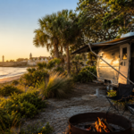 Luxury RV campsite overlooking the Atlantic Ocean near historic St. Augustine at sunset, featuring a polished travel trailer, campfire, palm trees, coastal dunes, and breathtaking views of the waterfront and historic skyline